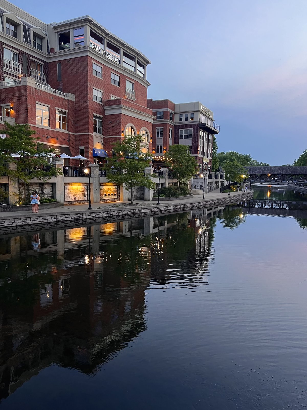 A brick riverfront at dusk, lights reflecting on still water, covered bridge beyond